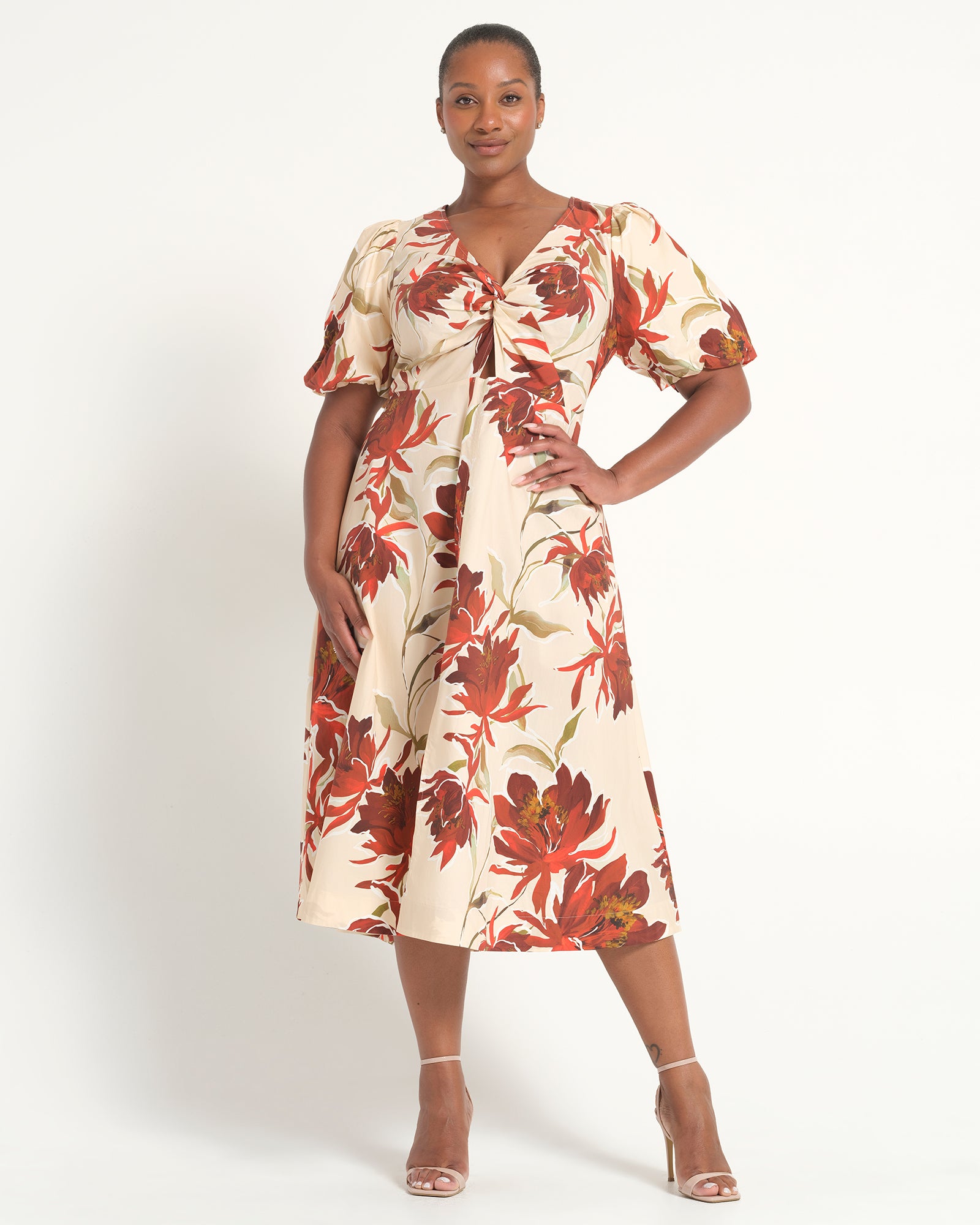 A woman modeling a cream-colored midi dress with a bold red and brown floral print, featuring puff sleeves and a gathered sweetheart neckline, set against a plain white studio background.