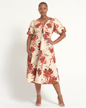 A woman modeling a cream-colored midi dress with a bold red and brown floral print, featuring puff sleeves and a gathered sweetheart neckline, set against a plain white studio background.