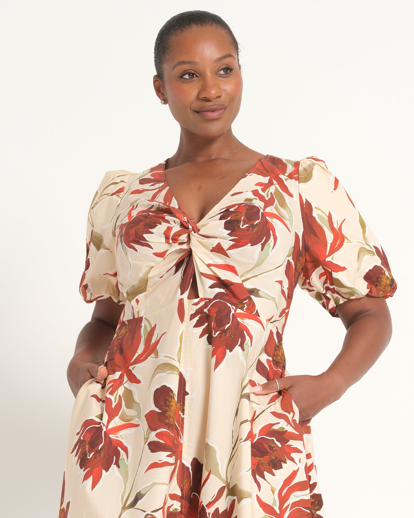 A woman modeling a short-sleeve cream-colored dress with a bold red and brown floral print, featuring a twisted front detail and puff sleeves, set against a plain white studio background.