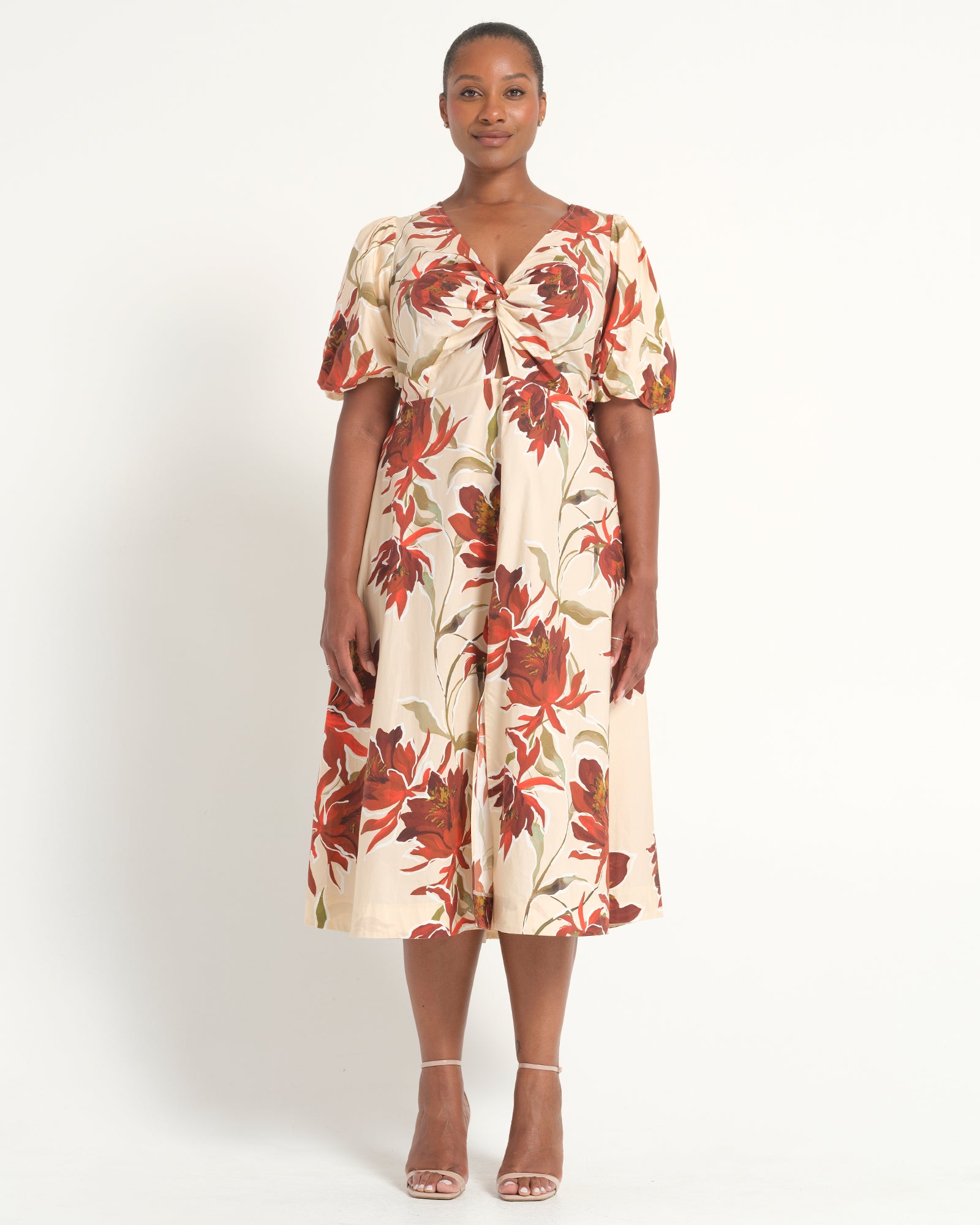 A woman modeling a short-sleeve midi dress with a cream base and a red, rust, and olive floral print, standing against a plain white studio background.