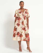 A woman modeling a cream-colored midi dress with a bold red and brown floral print, featuring puff sleeves and a gathered sweetheart neckline, set against a plain white studio background.