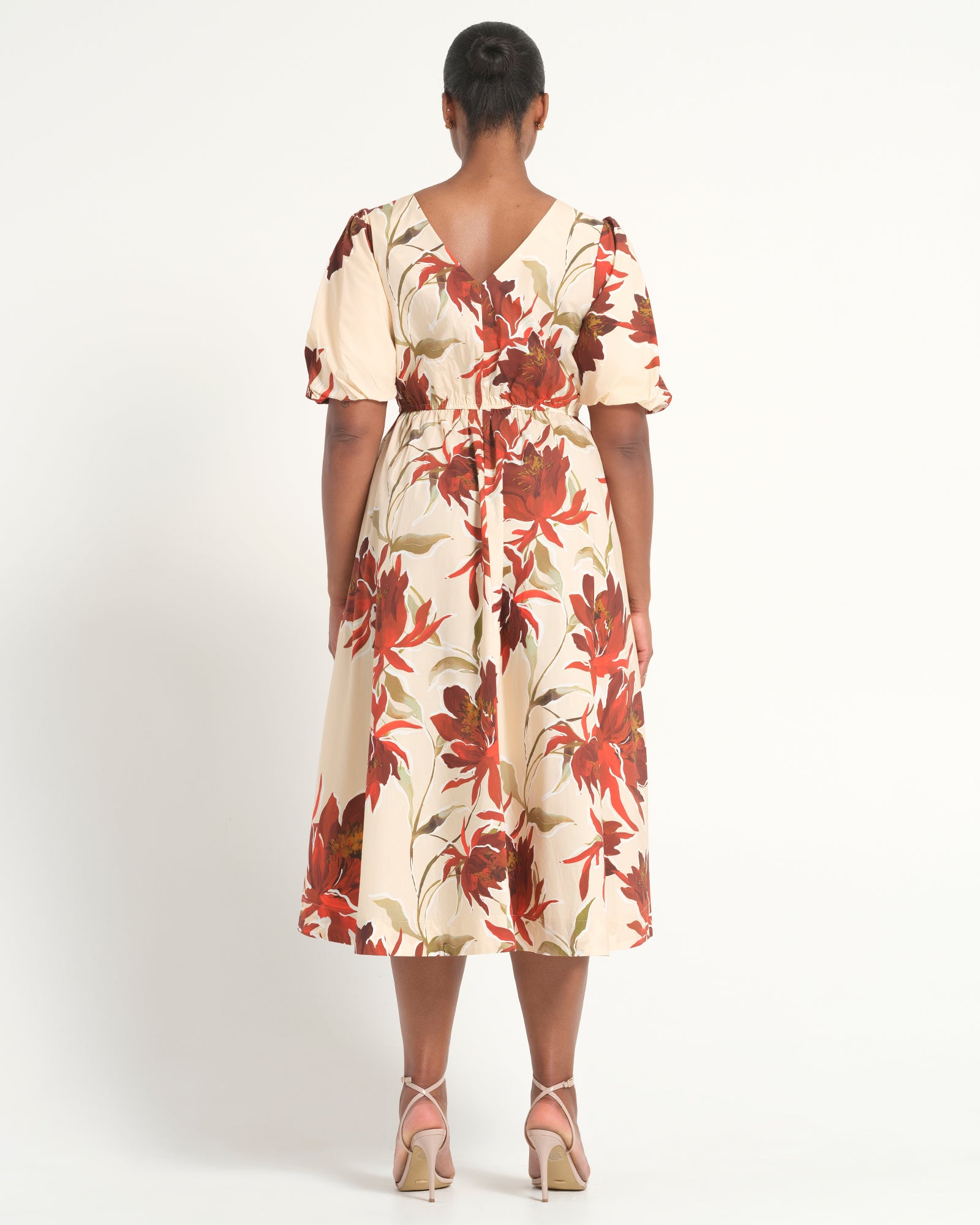 A woman viewed from the back wearing a cream midi dress with a red and brown floral print, short puff sleeves, and a tie waist, paired with light pink high-heeled sandals against a white studio background.