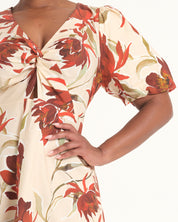 A close-up of a model wearing a cream-colored dress with a bold red and orange floral print and a twisted front detail.