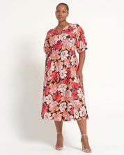A woman modeling a short-sleeved midi dress with a vibrant red, pink, and orange floral pattern on a black background, paired with clear high-heeled sandals against a plain white studio background.