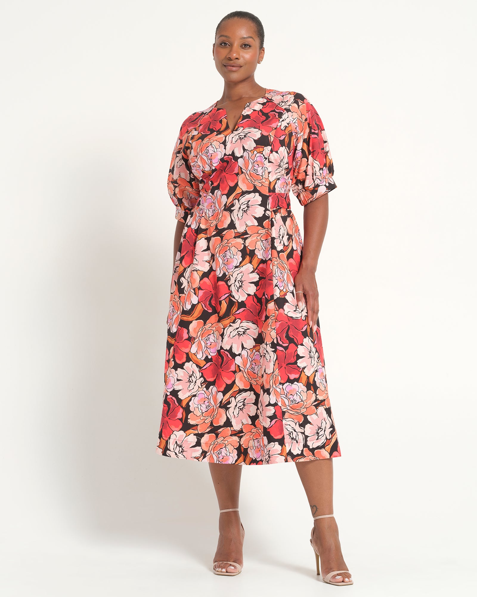 A woman modeling a short-sleeved midi dress with a vibrant red, pink, and orange floral pattern on a black background, paired with clear high-heeled sandals against a plain white studio background.