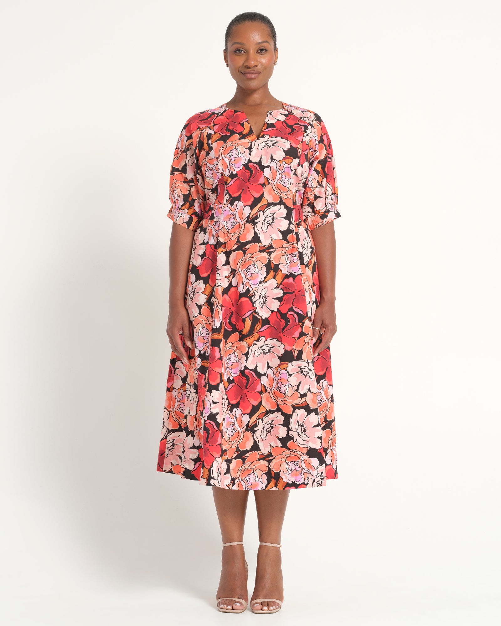 A woman modeling a short-sleeve midi dress with a vibrant red, pink, and orange floral print on a black background, paired with clear high-heeled sandals, against a plain white studio background.