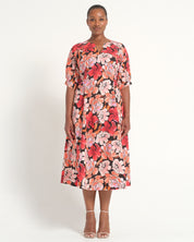 A woman modeling a short-sleeve midi dress with a vibrant red, pink, and orange floral print on a black background, paired with clear high-heeled sandals, against a plain white studio background.