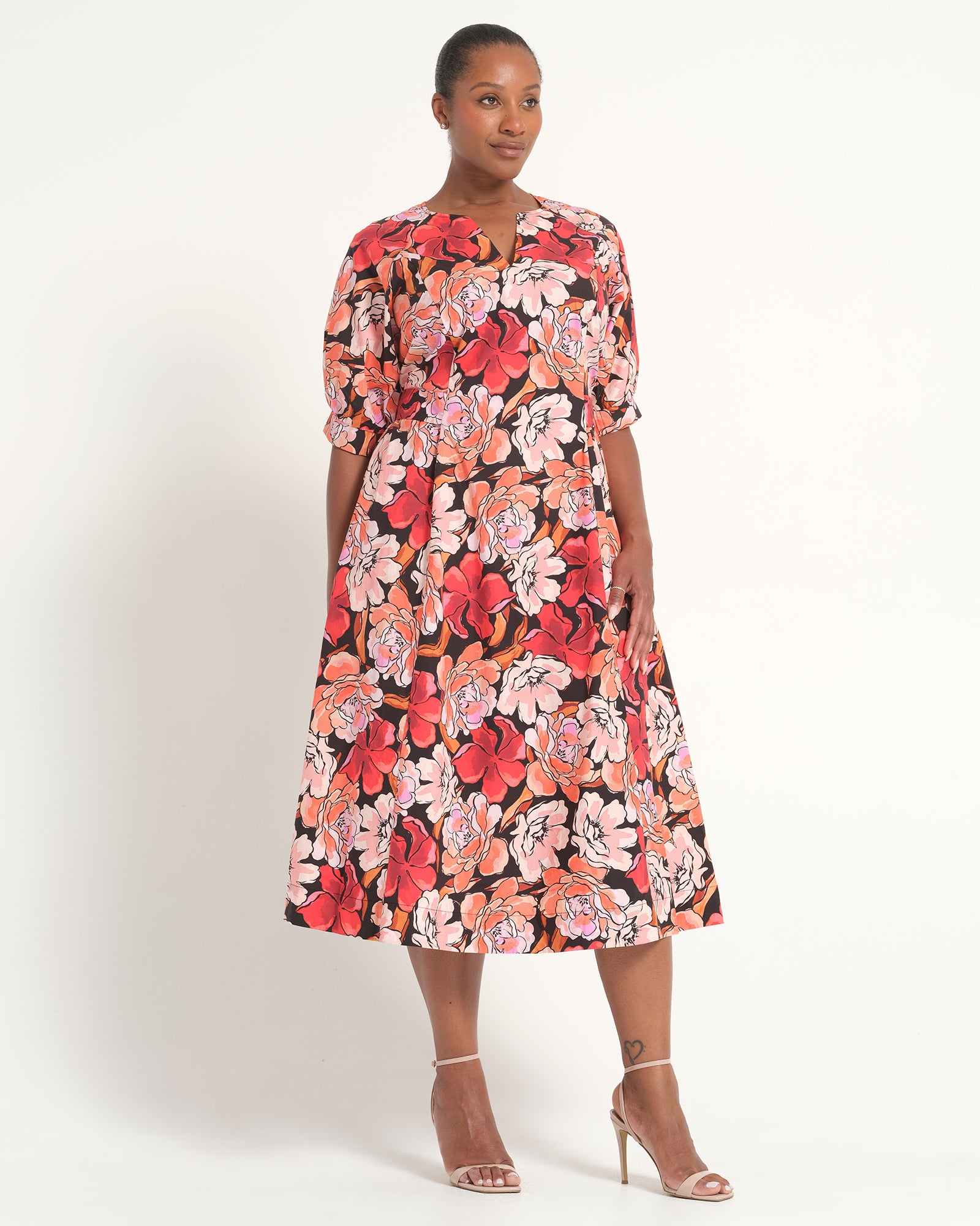 A woman modeling a knee-length floral dress with short sleeves, a split neckline, and a vibrant print of red, pink, and orange flowers on a black background, paired with clear high-heeled sandals against a plain white studio background.