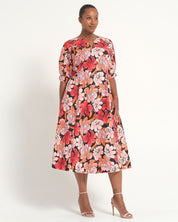 A woman modeling a knee-length floral dress with short sleeves, a split neckline, and a vibrant print of red, pink, and orange flowers on a black background, paired with clear high-heeled sandals against a plain white studio background.