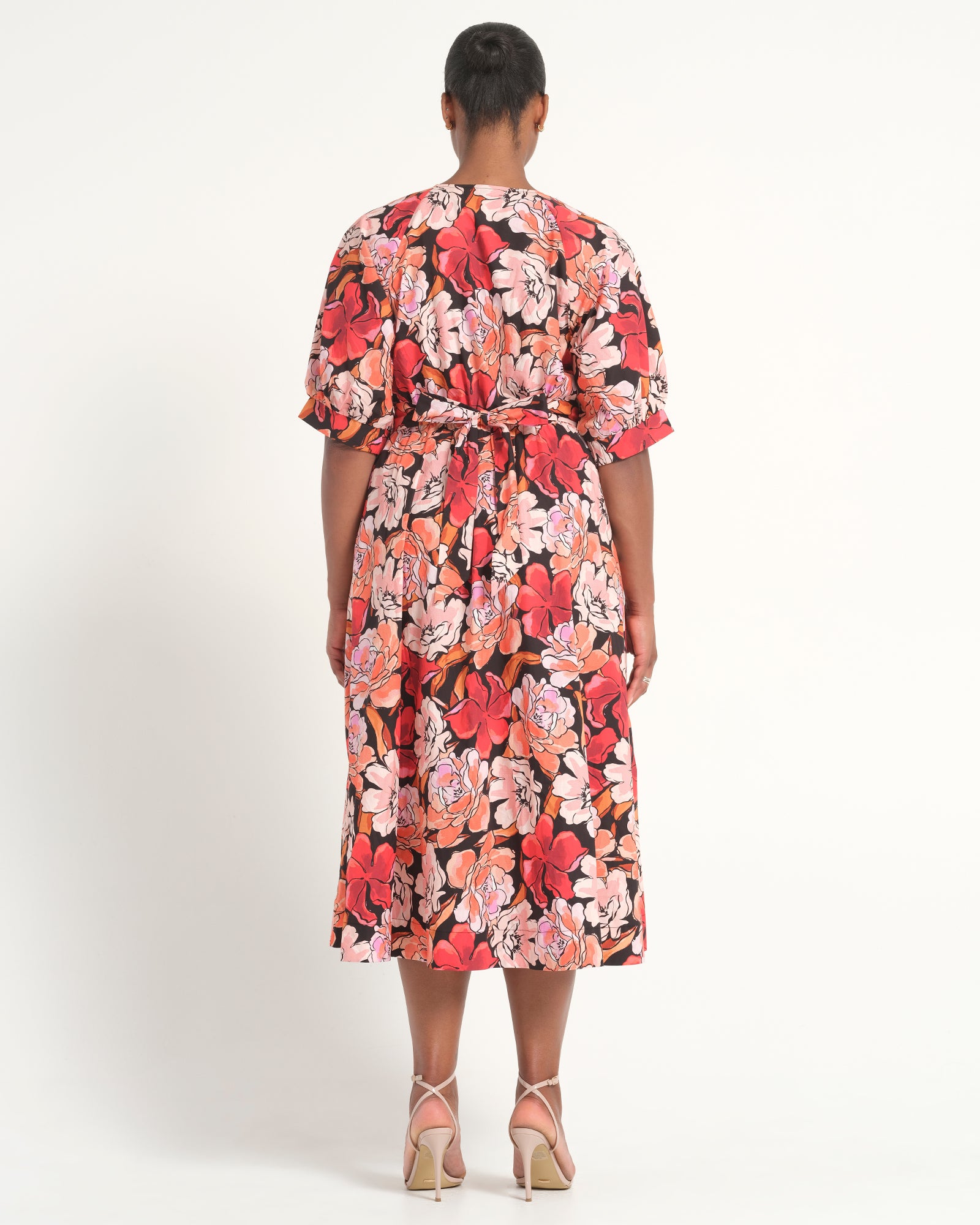 A woman viewed from the back wearing a short-sleeve midi dress with a vibrant red, pink, and orange floral print on a black background. She is wearing beige high-heeled sandals against a plain white studio background.