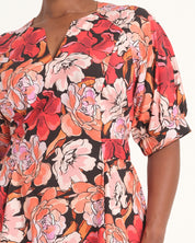 A close-up of a model wearing a short-sleeve wrap-style dress with a vibrant floral print in red, pink, orange, and white on a black background.