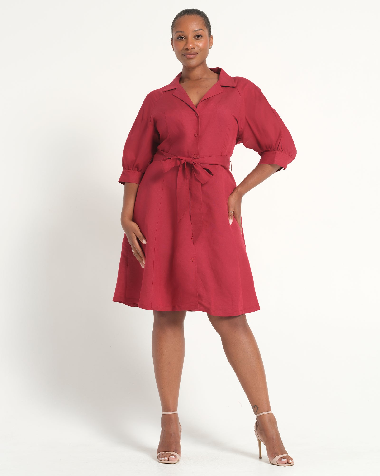 A woman modeling a red midi shirt dress with puff sleeves, a pointed collar, and a matching waist tie, photographed against a plain white studio background.