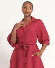 A woman wearing a red collared shirt dress with puff sleeves, a matching waist tie belt, and side pockets, photographed against a plain light background.