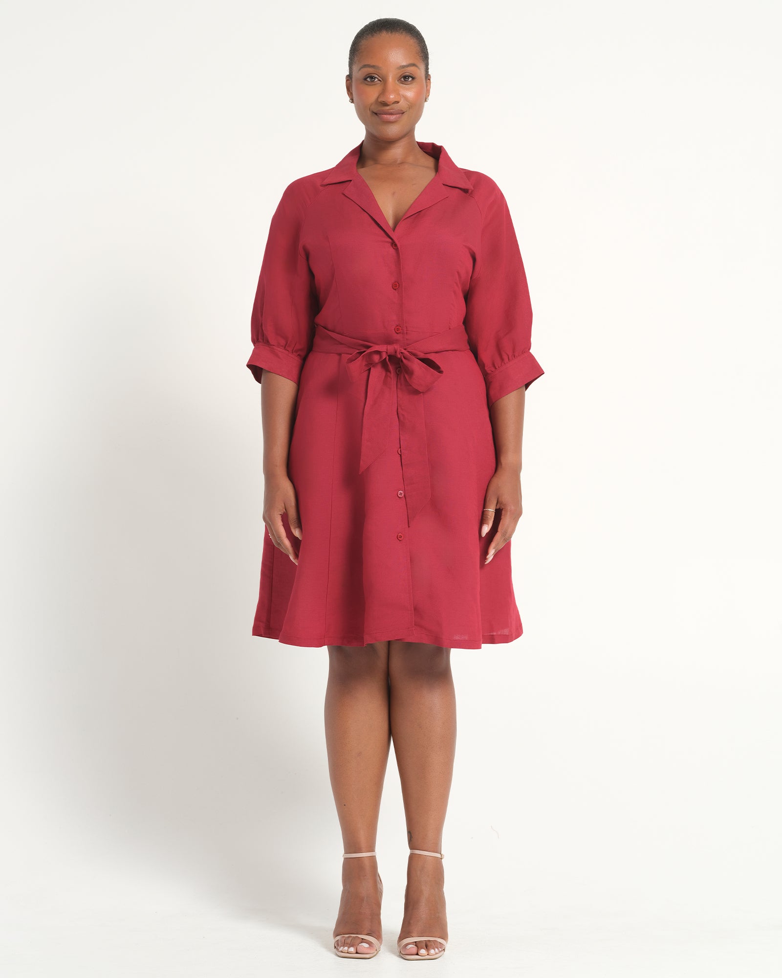 A woman modeling a red collared shirt dress with three-quarter sleeves, rolled cuffs, and a matching tie belt at the waist, photographed against a plain light gray studio background.