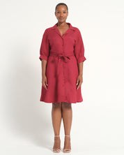 A woman modeling a red collared shirt dress with three-quarter sleeves, rolled cuffs, and a matching tie belt at the waist, photographed against a plain light gray studio background.