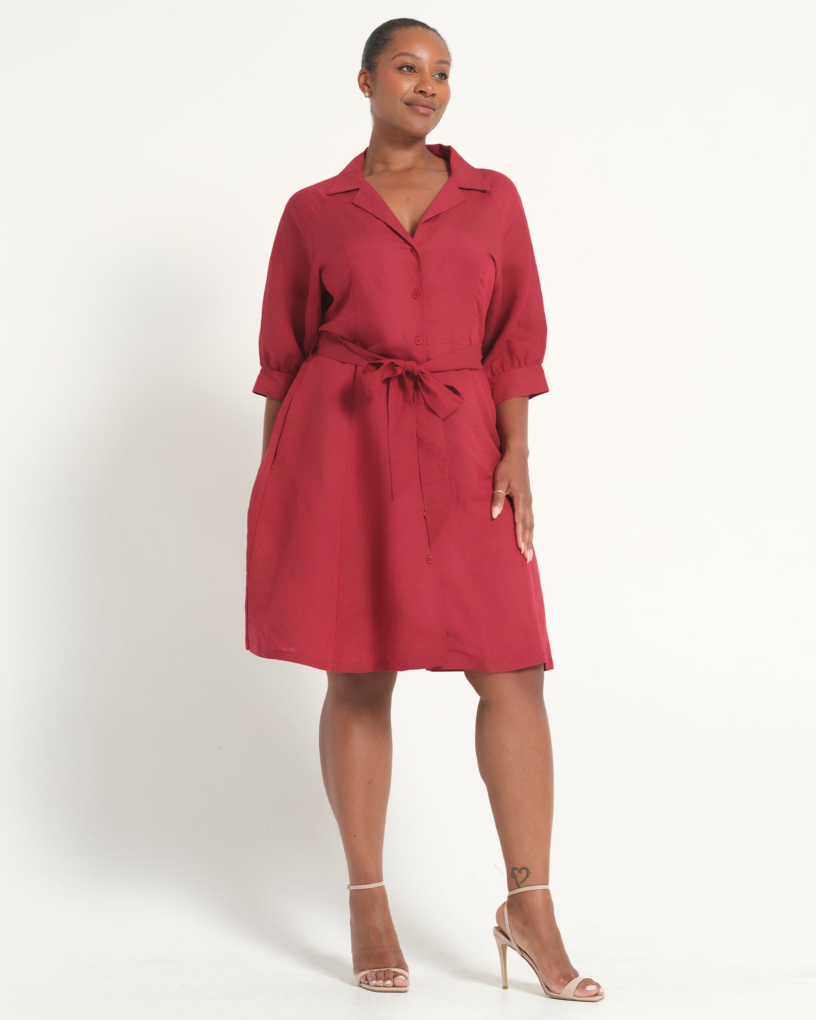 A woman modeling a red collared shirt dress with three-quarter sleeves, a matching tie belt, and side pockets, paired with clear high-heeled sandals against a plain white studio background.