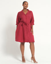 A woman modeling a red collared shirt dress with three-quarter sleeves, a matching tie belt, and side pockets, paired with clear high-heeled sandals against a plain white studio background.
