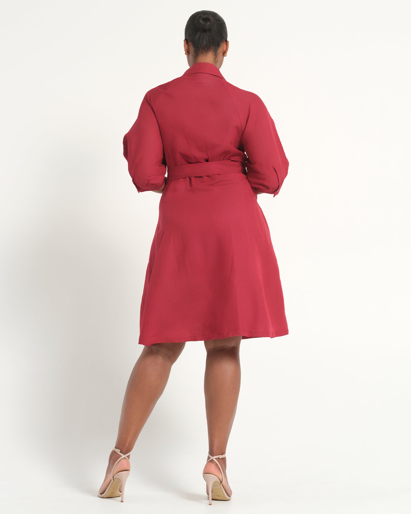 A woman viewed from the back wearing a red belted wrap dress with long sleeves, paired with beige high-heeled sandals, against a plain white studio background.