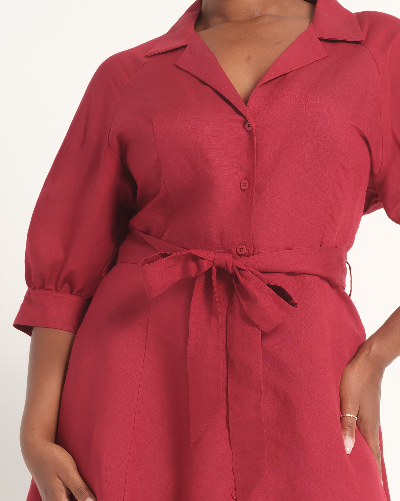 A close-up of a model wearing a red collared shirt dress with three-quarter sleeves, a button-down front, and a matching fabric tie belt at the waist.