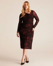 A woman models a black knee-length dress with a red leafy floral pattern, long sheer sleeves, and a draped crossover neckline, paired with black pointed-toe heels against a solid light beige background.