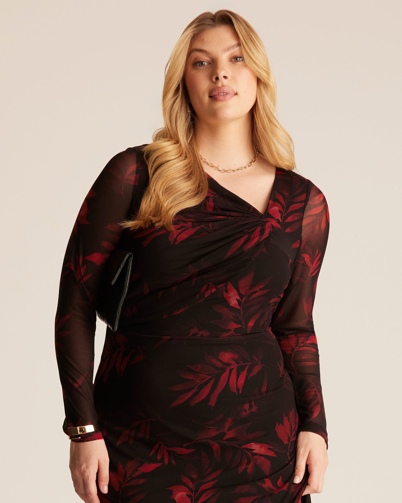 A plus-size blonde model wearing a black long-sleeve wrap dress with a red tropical leaf pattern, posed against a solid beige studio background.