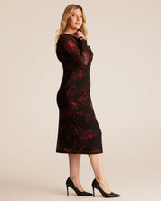 A woman modeling a long-sleeve midi dress with a black base and red floral pattern, paired with black high heels, against a solid light beige studio background.