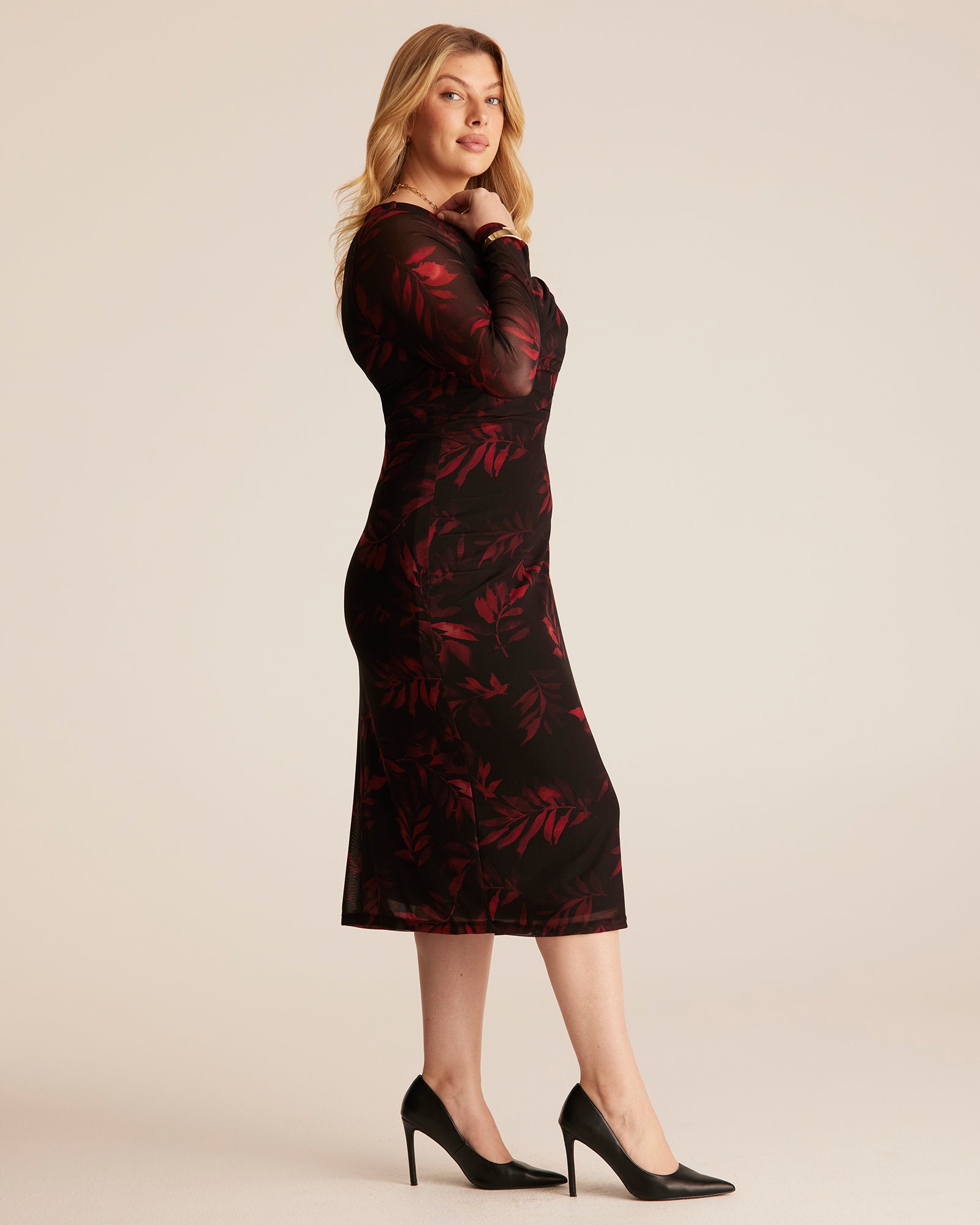 A woman modeling a long-sleeve midi dress with a black base and red floral pattern, paired with black high heels, against a solid light beige studio background.