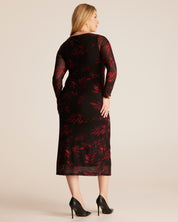 A woman shown from the back wearing a black midi dress with a red floral pattern and sheer long sleeves, paired with black high heels, against a solid light beige background.
