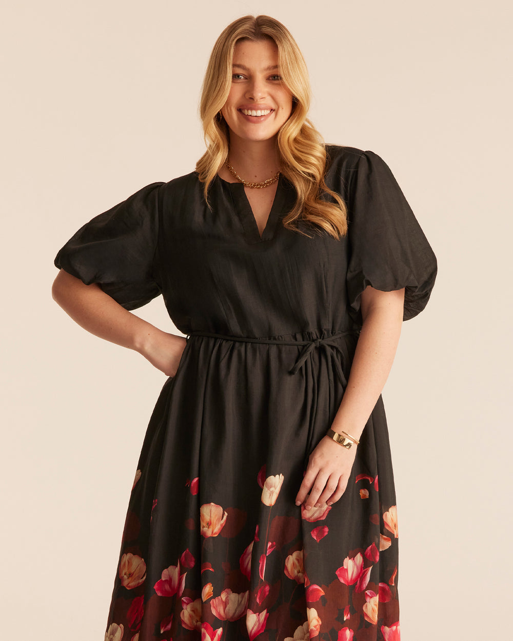A woman modeling a black midi dress with short puff sleeves, a V-neckline, and a red and beige tulip print concentrated along the lower skirt of a black dress, photographed against a plain beige studio background.