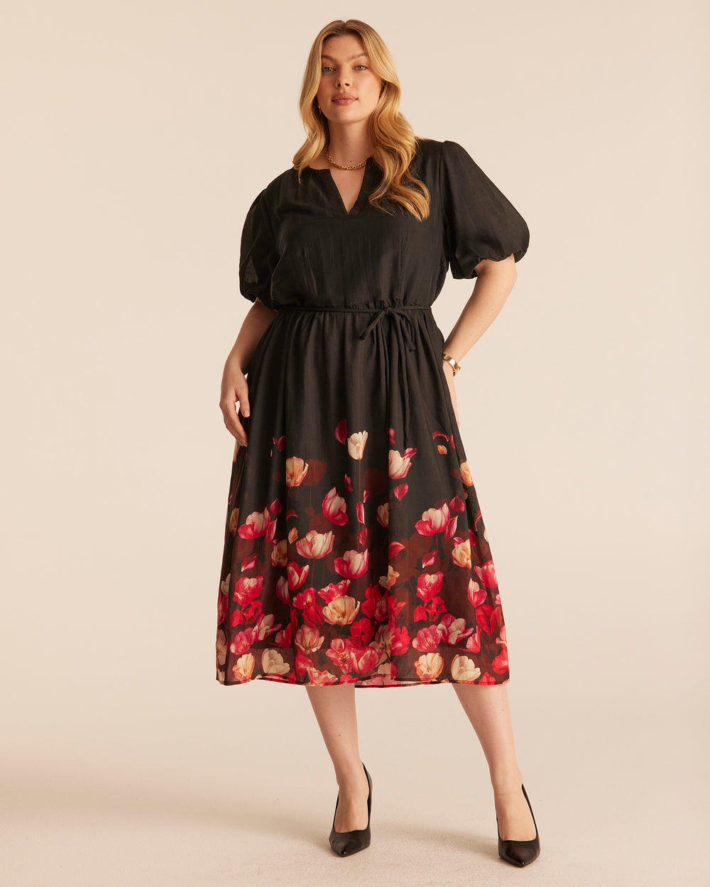 A woman modeling a black midi dress with short puff sleeves, a V-neckline, and a red and beige tulip print concentrated along the lower skirt of a black dress, photographed against a plain beige studio background.