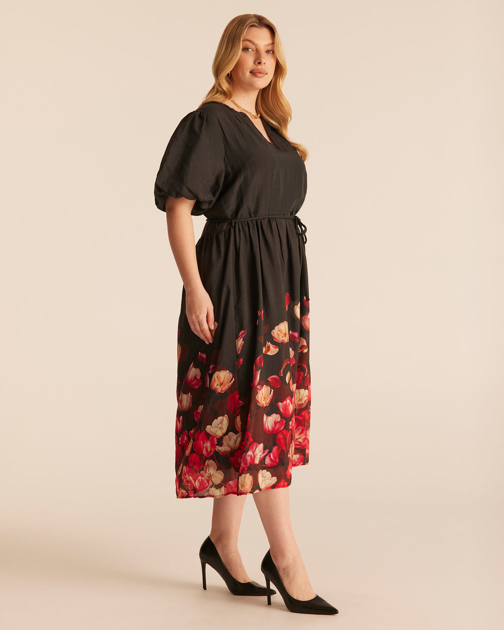 A woman modeling a black midi dress with short puff sleeves, a V-neckline, and a red and beige tulip print concentrated along the lower skirt of a black dress, photographed against a plain beige studio background.