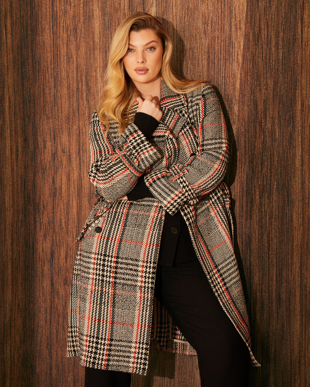 Woman wearing a just below the knee, double breasted coat. Its colour consists of a beige base and black and tangerine orange plaid pattern. The model stands against a beige background.