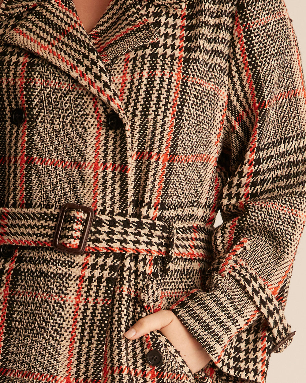 Woman wearing a just below the knee, double breasted coat. Its colour consists of a beige base and black and tangerine orange plaid pattern. The model stands against a beige background.