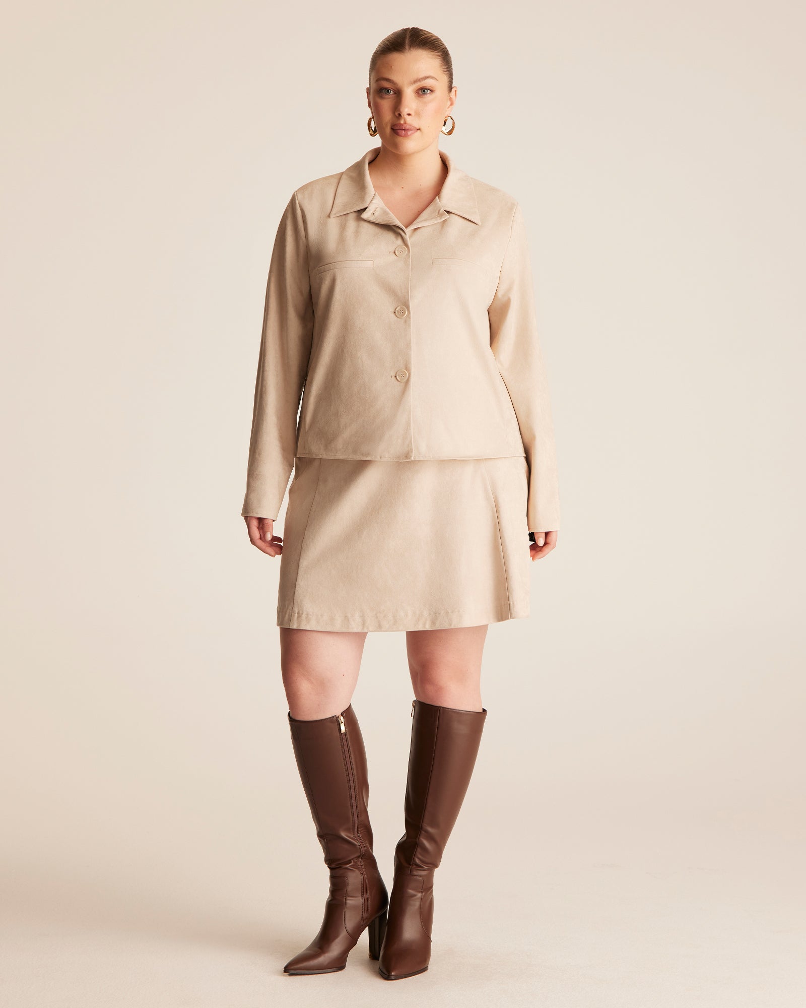 Woman wearing a beige jacket and skirt set with brown knee-high boots on a plain background