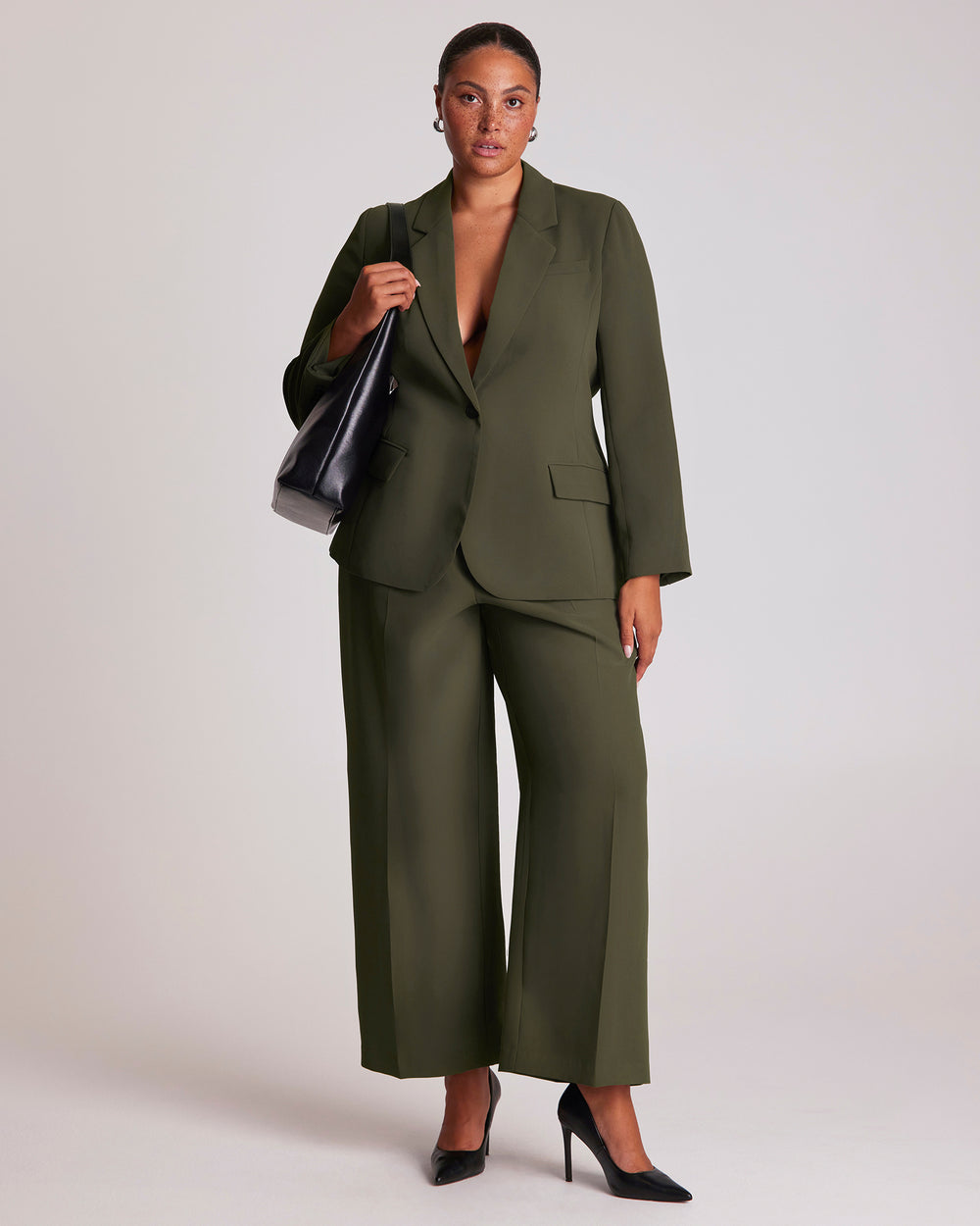 Woman wearing a dark green suit blazer, holding a black bag against a plain background.