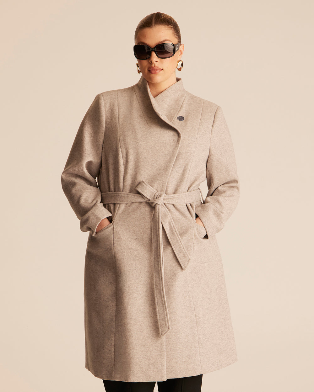 Woman wearing a beige trench style coat with an asymmetrical funnel style neckline on a beige background with black tailored pants.