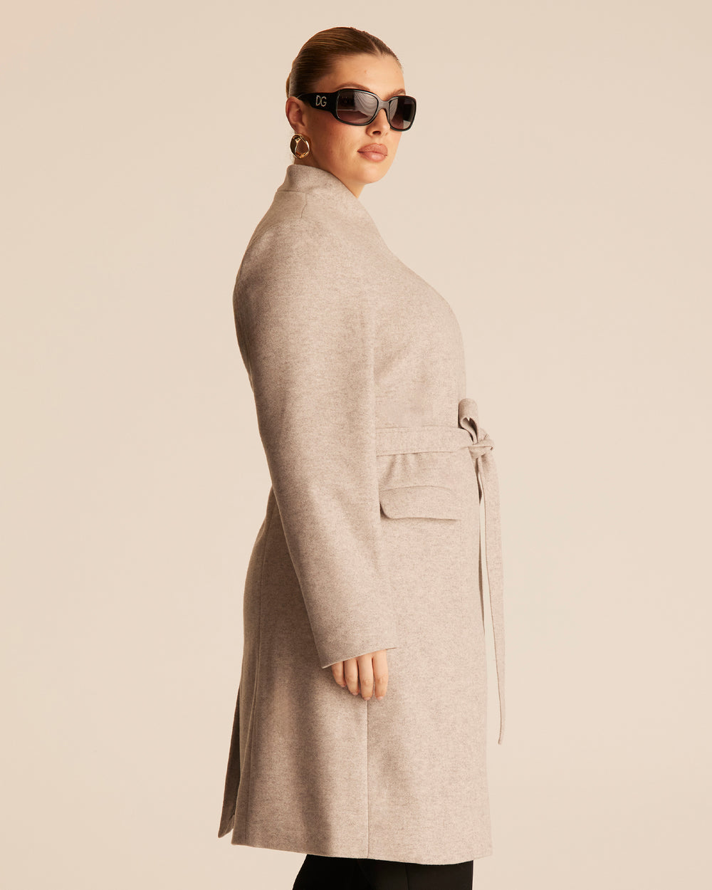 Woman wearing a beige trench style coat with an asymmetrical funnel style neckline on a beige background with black tailored pants.