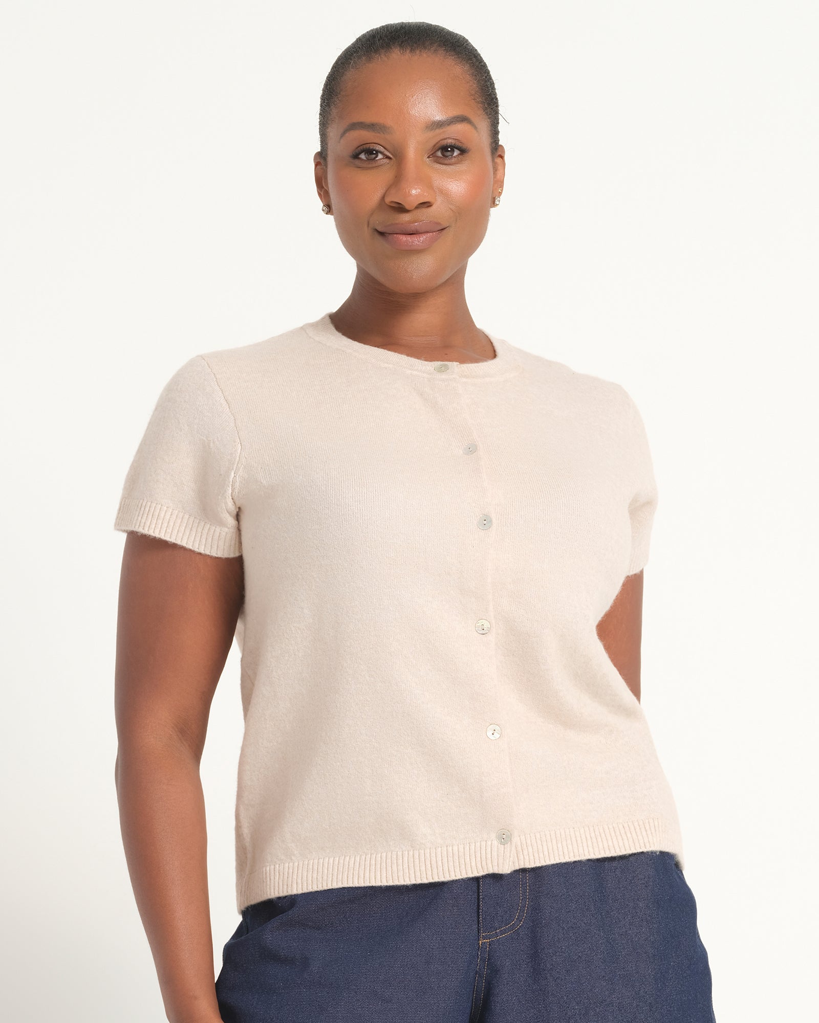 A woman wearing a short-sleeve cream-colored knit cardigan with a button-up front, paired with dark blue jeans, against a plain white studio background.