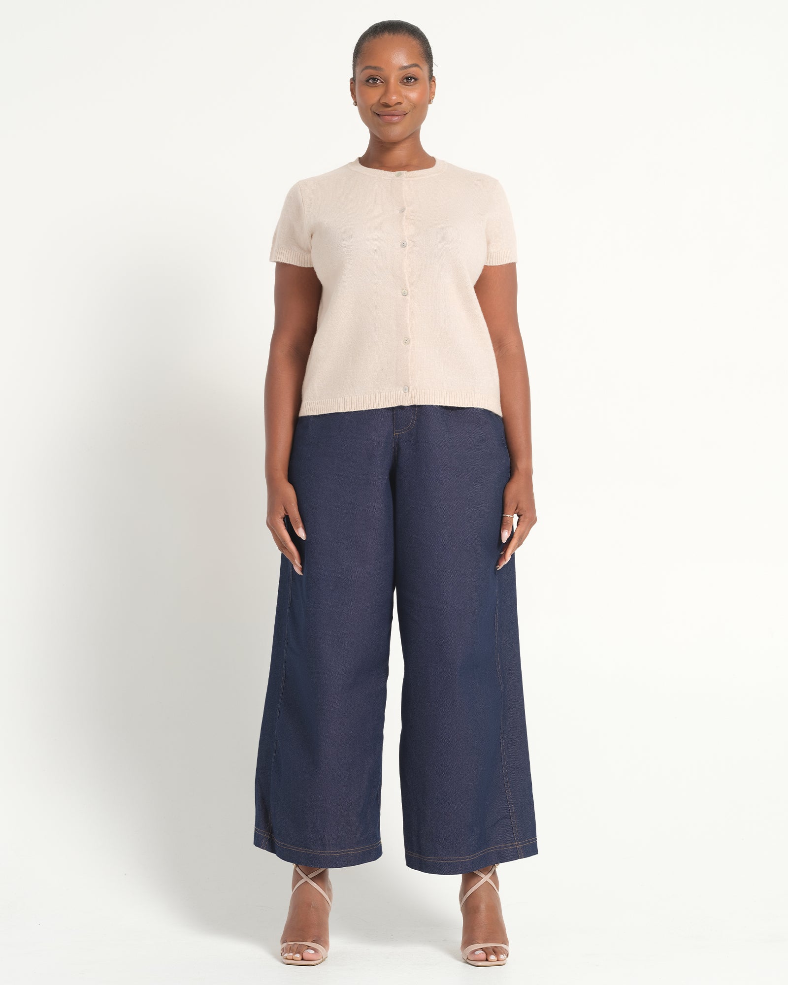 A woman modeling a cream short-sleeve button-front top paired with navy wide-leg cropped pants and nude strappy heels, photographed against a clean off-white studio background.