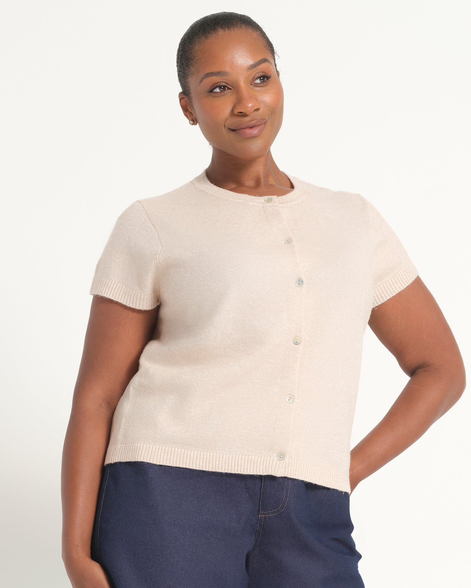 A woman modeling a short-sleeve knit cardigan in a light cream color with a button-up front, paired with navy blue pants, against a plain white studio background.