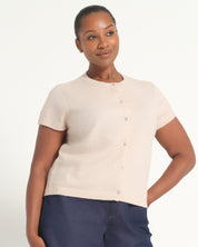 A woman modeling a short-sleeve knit cardigan in a light cream color with a button-up front, paired with navy blue pants, against a plain white studio background.