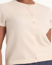 A close-up of a person wearing a short-sleeve knit cardigan top in a light cream or off-white color, with small buttons down the front. The background is a clean white studio setting.