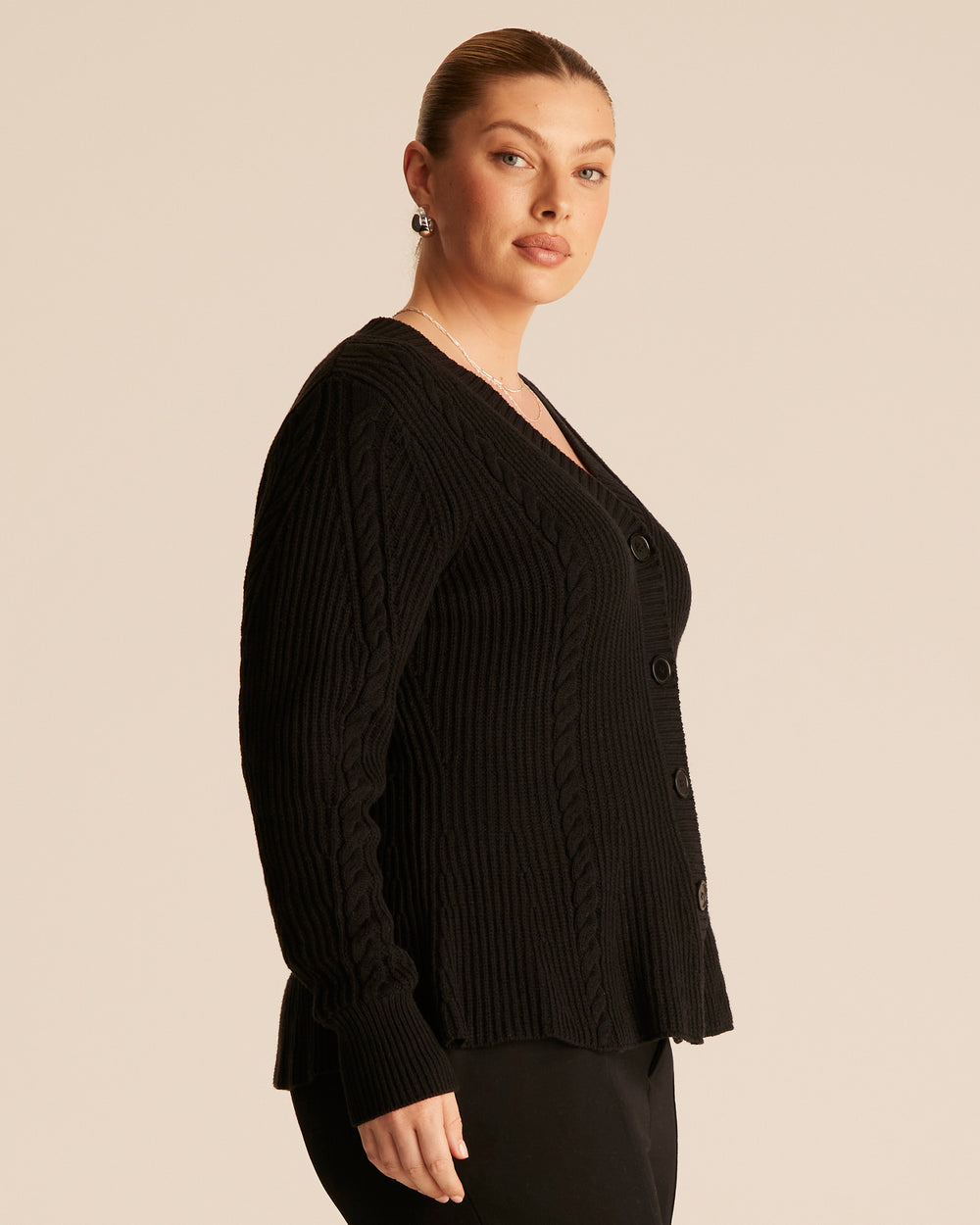 Woman wearing a black ribbed cable knit cardigan with a button up front and V-neck design against a beige background