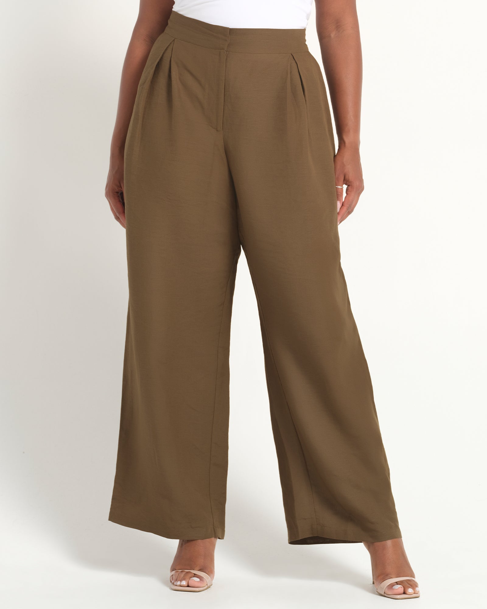 A model wearing high-waisted, wide-leg pleated trousers in a solid taupe-brown color, paired with a white top and clear high-heeled sandals, against a plain white studio background.