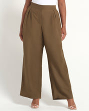 A model wearing high-waisted, wide-leg pleated trousers in a solid taupe-brown color, paired with a white top and clear high-heeled sandals, against a plain white studio background.