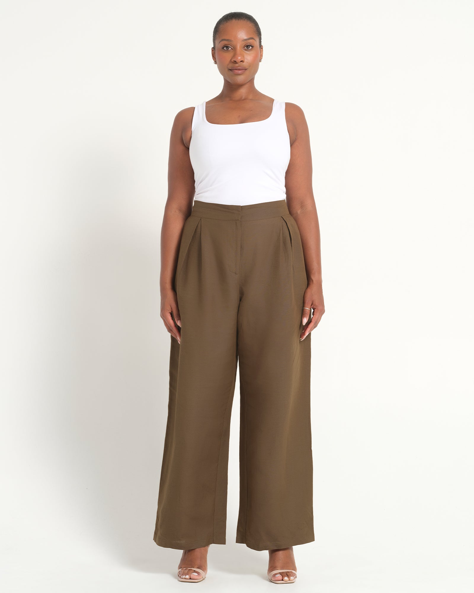 A woman modeling a pair of high-waisted, wide-leg brown trousers paired with a white tank top, standing against a plain off-white studio background.