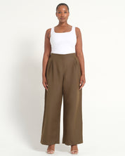A woman modeling a pair of high-waisted, wide-leg brown trousers paired with a white tank top, standing against a plain off-white studio background.