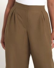 A close-up of a person wearing high-waisted, wide-leg brown trousers with pleated detailing at the front, paired with a white top.