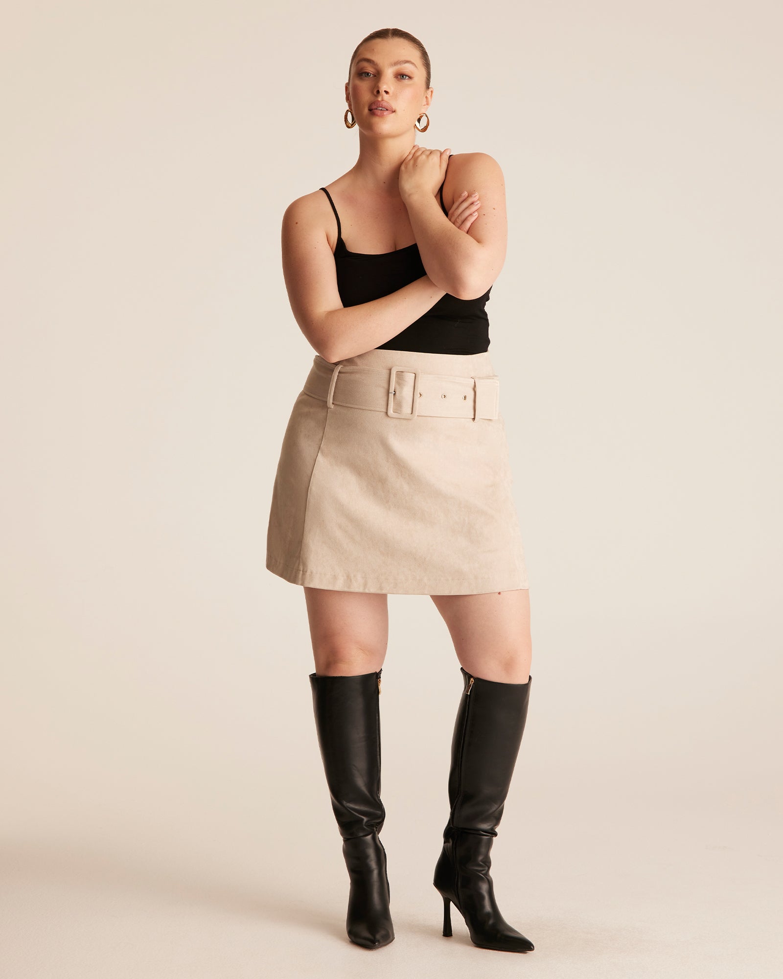 Woman wearing a black top and beige skirt with black knee-high boots on a plain background