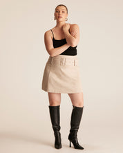 Woman wearing a black top and beige skirt with black knee-high boots on a plain background